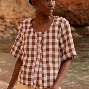 May include: A brown and white gingham button-up shirt with short sleeves. The shirt has a square neckline and a relaxed fit.