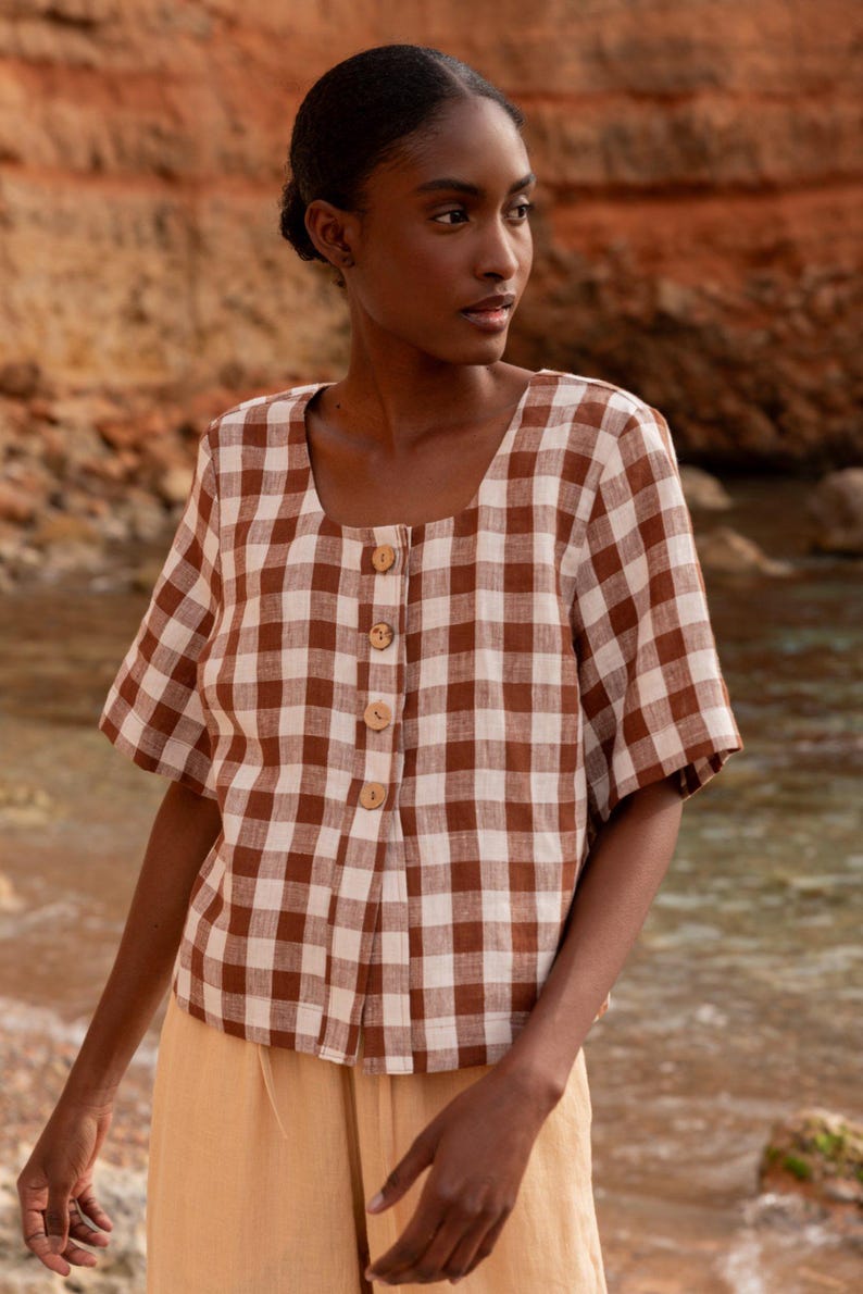 May include: A brown and white gingham button-up shirt with short sleeves. The shirt has a square neckline and a relaxed fit.