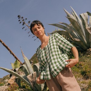 May include: A woman wearing a green and white gingham button-up shirt with brown linen pants stands in a field of agave plants.