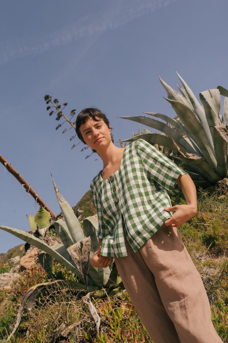 May include: A woman wearing a green and white gingham button-up shirt with brown linen pants stands in a field of agave plants.