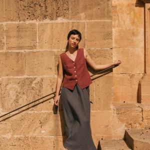 May include: A woman wearing a burgundy sleeveless linen vest with a button closure and a dark gray linen skirt. She is standing on a set of stone steps in front of a stone wall.