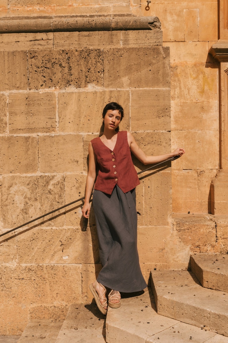 May include: A woman wearing a burgundy sleeveless linen vest with a button closure and a dark gray linen skirt. She is standing on a set of stone steps in front of a stone wall.