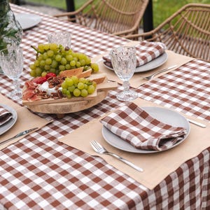 May include: A brown and white checkered tablecloth with a wooden serving board with grapes, cheese, and prosciutto. There are two place settings with brown and white checkered napkins, plates, and silverware.