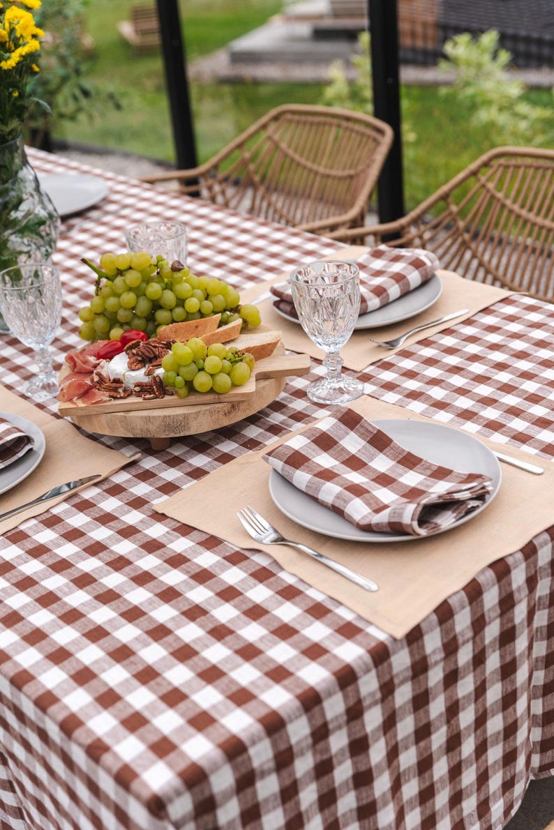 May include: A brown and white checkered tablecloth with a wooden serving board with grapes, cheese, and prosciutto. There are two place settings with brown and white checkered napkins, plates, and silverware.