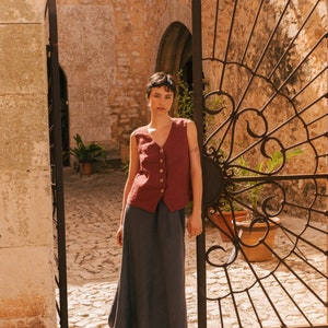 May include: A woman wearing a maroon sleeveless linen vest with button closures and a dark gray maxi skirt. She is standing in front of a wrought iron gate.