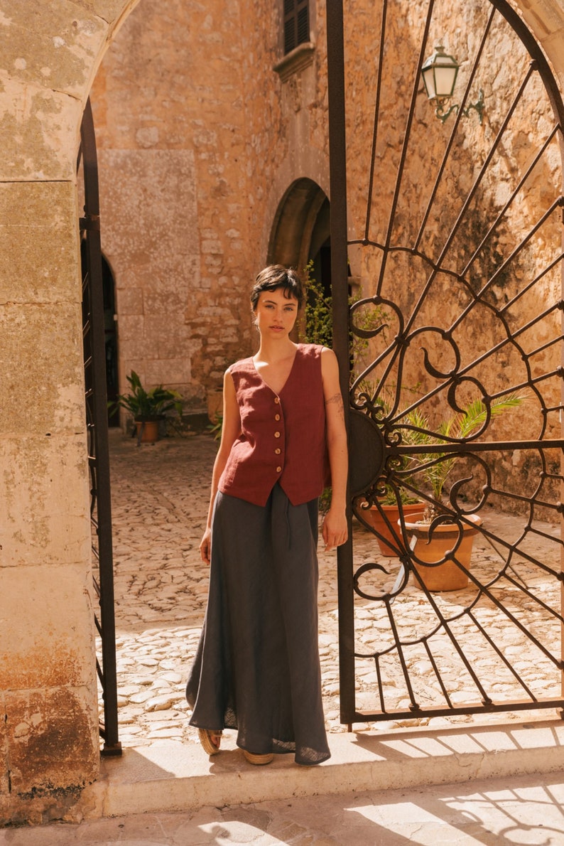 May include: A woman wearing a maroon sleeveless linen vest with button closures and a dark gray maxi skirt. She is standing in front of a wrought iron gate.