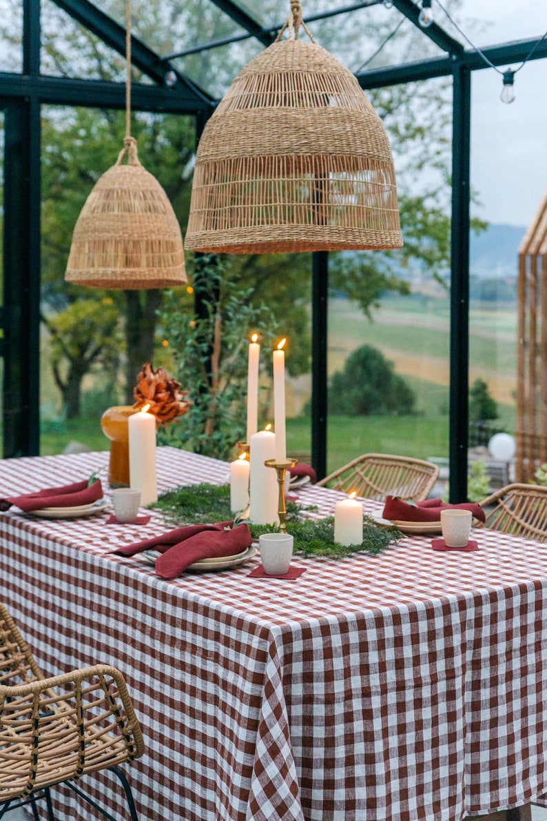 May include: A red and white checkered tablecloth is set on a table in a greenhouse. The table is decorated with candles, greenery, and wicker chairs.