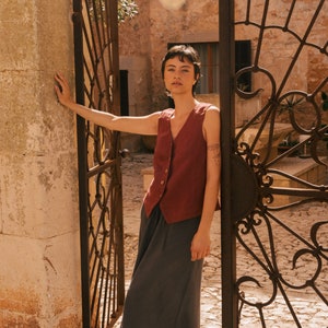 May include: A woman wearing a rust-colored linen vest with a button closure over a dark gray maxi skirt. She is standing in front of a wrought iron gate.