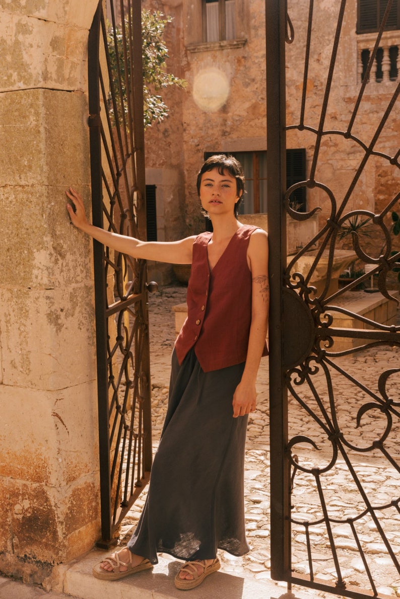 May include: A woman wearing a rust-colored linen vest with a button closure over a dark gray maxi skirt. She is standing in front of a wrought iron gate.