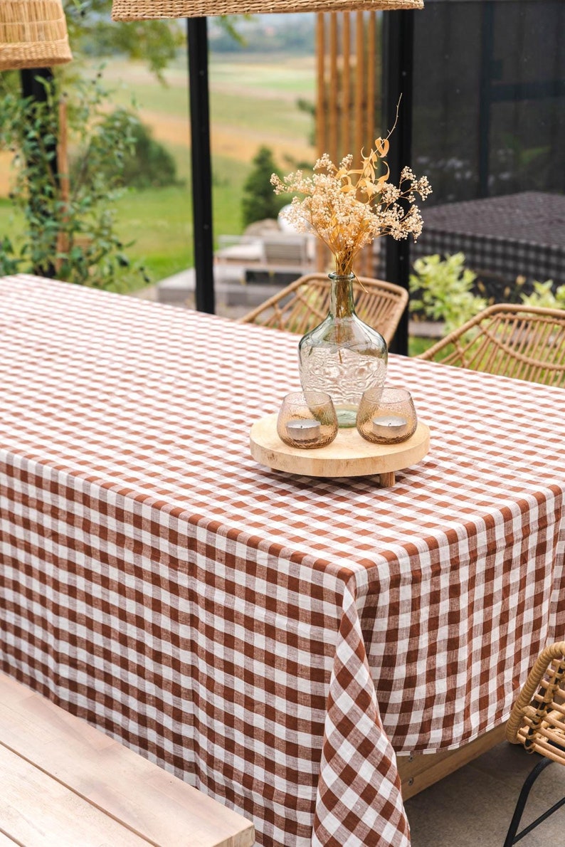 May include: A brown and white checkered tablecloth covers a wooden table with a glass vase filled with dried flowers and two glass candle holders on a wooden tray.