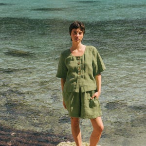 May include: A woman wearing a green linen short-sleeved top and matching shorts stands on a rock by the water. She is wearing brown sandals.