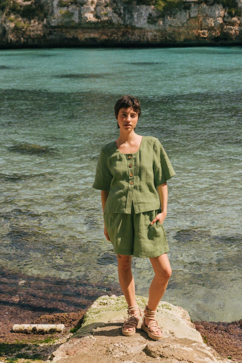 May include: A woman wearing a green linen short-sleeved top and matching shorts stands on a rock by the water. She is wearing brown sandals.