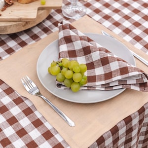 May include: A brown and white checkered linen tablecloth with a beige placemat, a white plate, a brown and white checkered linen napkin, a fork, and a bunch of green grapes.