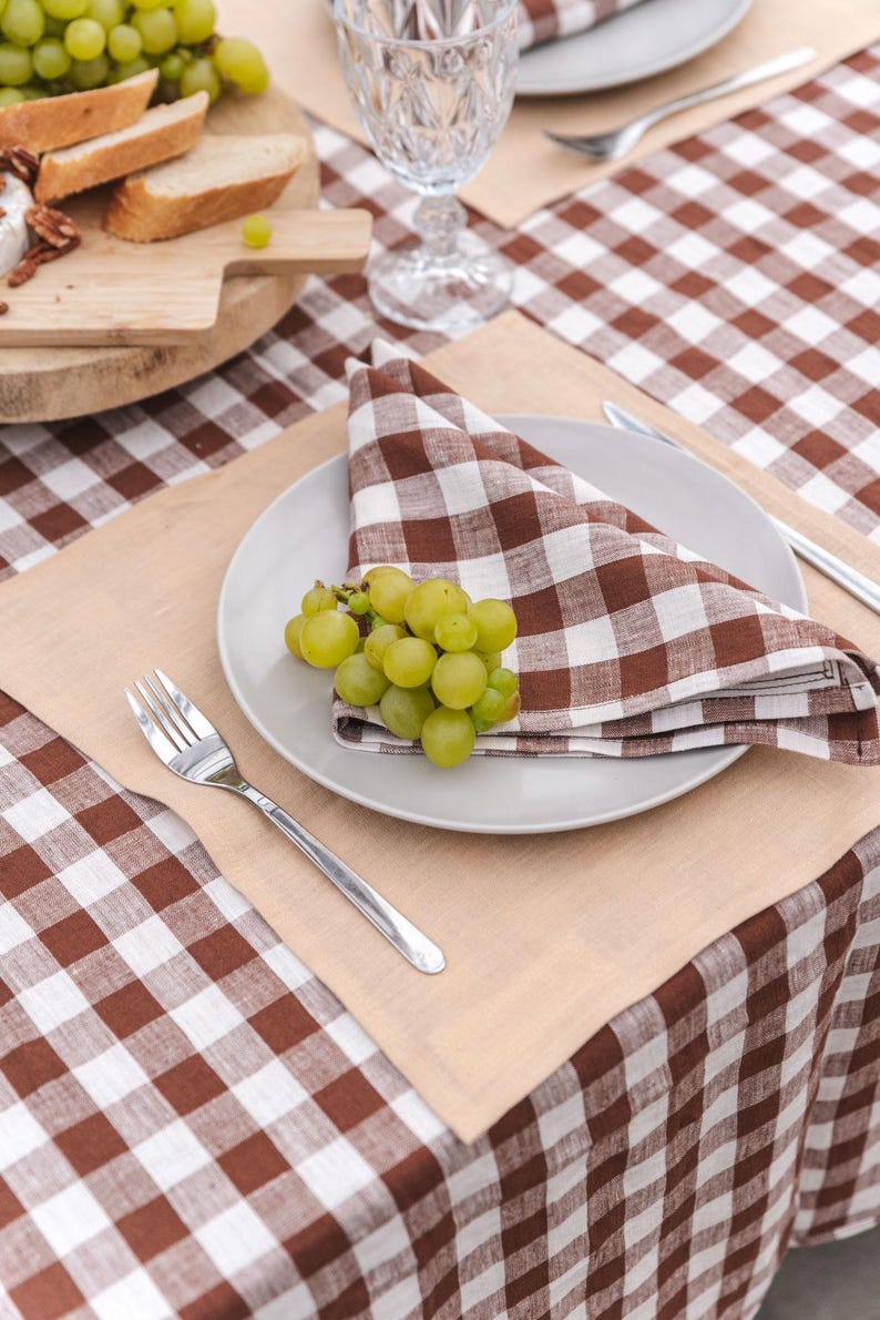 May include: A brown and white checkered linen tablecloth with a beige placemat, a white plate, a brown and white checkered linen napkin, a fork, and a bunch of green grapes.