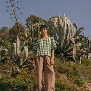 May include: A person wearing a green and white gingham button-up shirt with brown wide-leg pants. The person is standing on a rocky surface with green plants in the background.