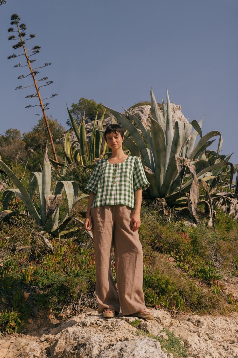 May include: A person wearing a green and white gingham button-up shirt with brown wide-leg pants. The person is standing on a rocky surface with green plants in the background.