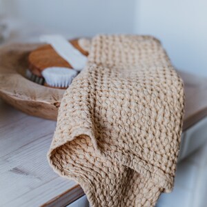 Op de afbeelding: Een beige wafeldoek gedrapeerd over een houten kom met een witte borstel erin. De handdoek is zacht en textuurrijk, en de kom is gemaakt van natuurlijk hout.