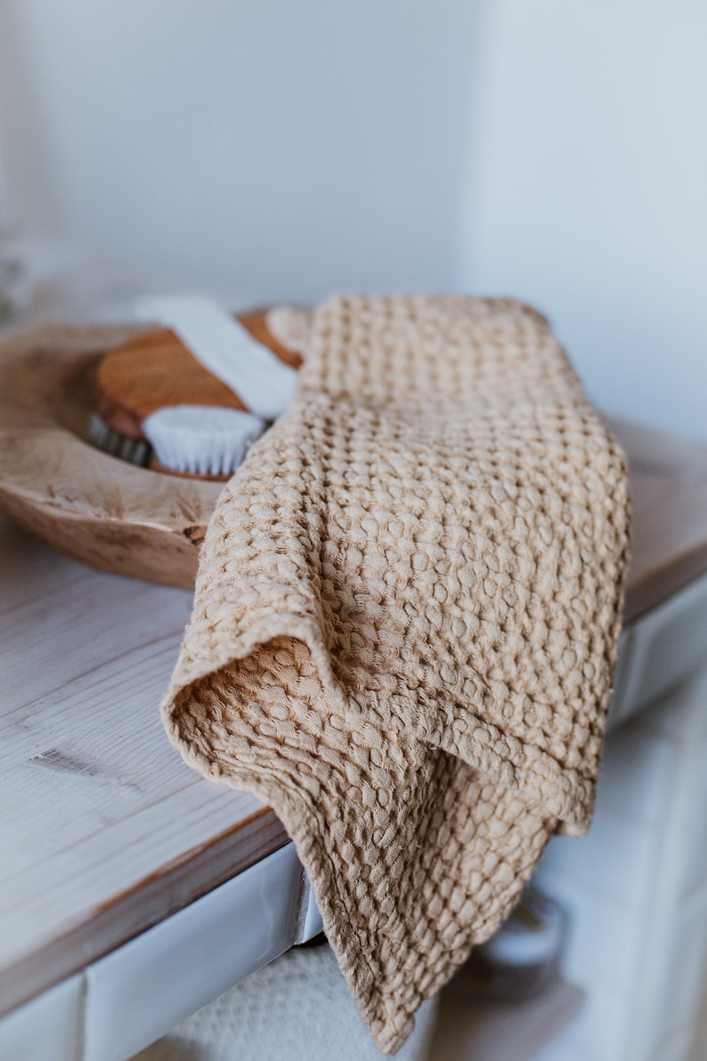 Op de afbeelding: Een beige wafeldoek gedrapeerd over een houten kom met een witte borstel erin. De handdoek is zacht en textuurrijk, en de kom is gemaakt van natuurlijk hout.