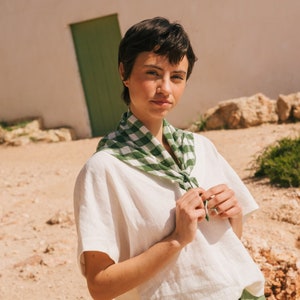May include: A person wearing a white linen shirt with a green and white checkered scarf tied around their neck. They are wearing green pants and standing in a desert landscape.