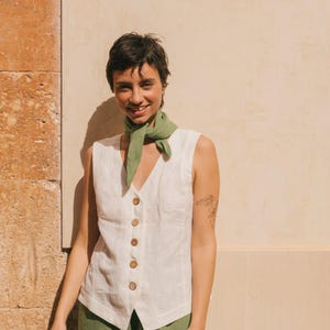May include: A woman wearing a white linen sleeveless top with a button-up front and a green scarf around her neck. She is wearing green linen pants and is standing in front of a tan wall.