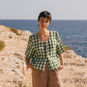 May include: A woman wearing a green and white gingham button-up shirt with brown linen pants standing on a rocky cliff overlooking the ocean.