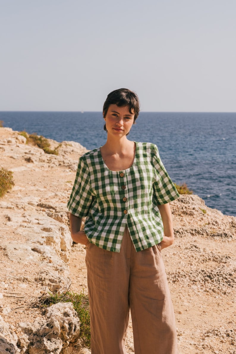 May include: A woman wearing a green and white gingham button-up shirt with brown linen pants standing on a rocky cliff overlooking the ocean.