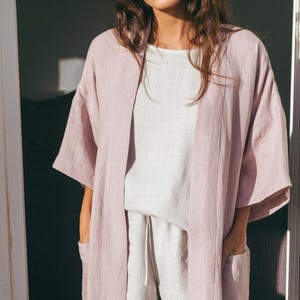 Peut inclure: Une longue robe en lin rose clair à la coupe ample et aux manches trois-quarts. La robe a un design simple avec un col châle et des poches sur les côtés.