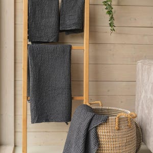 Op de afbeelding: Een houten ladder met drie grijze wafeldoeken die eraan hangen. Een grote geweven mand met touwhandvatten staat op de vloer met een grijze wafeldoek eroverheen gelegd.