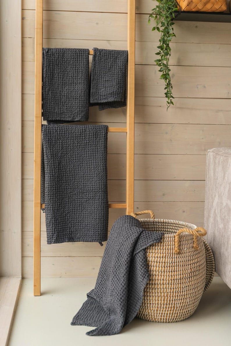 Op de afbeelding: Een houten ladder met drie grijze wafeldoeken die eraan hangen. Een grote geweven mand met touwhandvatten staat op de vloer met een grijze wafeldoek eroverheen gelegd.