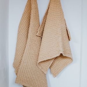 Op de afbeelding: Twee keukenhanddoeken met een wafelstructuur hangen aan een houten rek met houten knoppen. De handdoeken zijn lichtbeige.