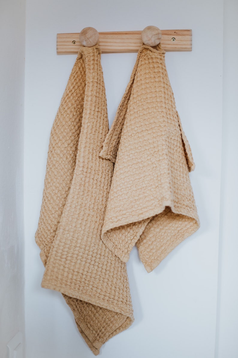 Op de afbeelding: Twee keukenhanddoeken met een wafelstructuur hangen aan een houten rek met houten knoppen. De handdoeken zijn lichtbeige.