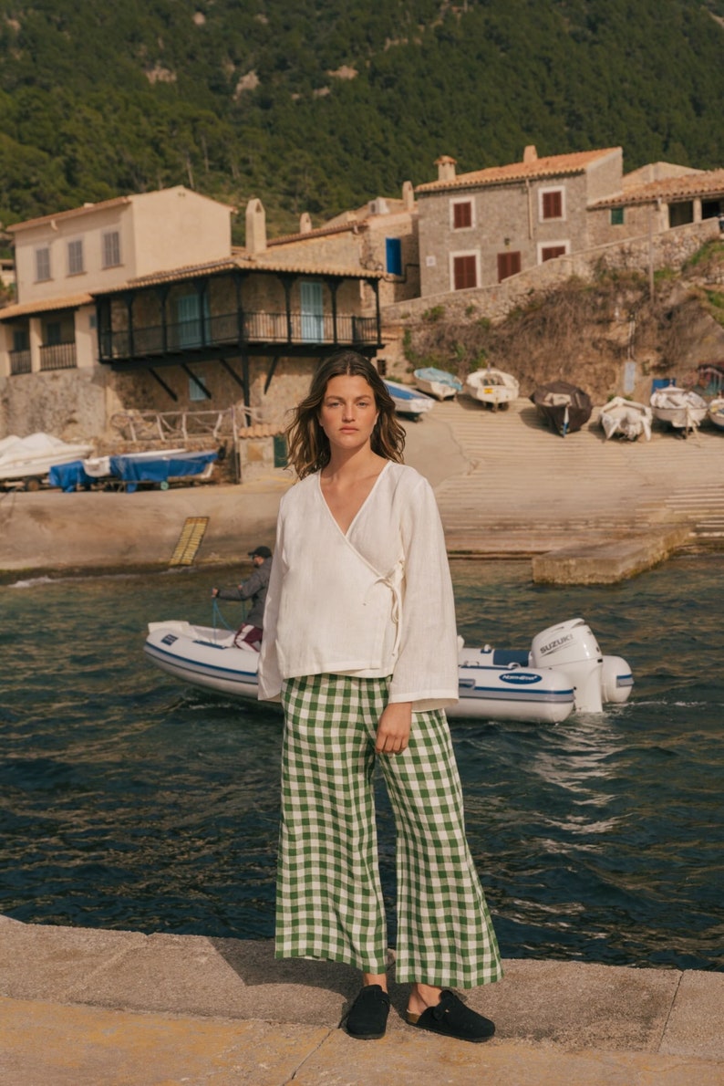 K&ouml;nnte beinhalten: Eine Frau in einem wei&szlig;en Leinen-Wickeltop und einer weiten gr&uuml;nen und wei&szlig;en Gingham-Hose steht auf einem Steg am Wasser. Sie blickt in die Kamera. Im Hintergrund sind Boote und ein kleines Dorf zu sehen.