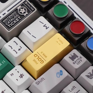 May include: A white mechanical keyboard with custom keycaps. The keycaps feature various colors, symbols, and text, including "NINTENDO CATALOG TICKET" and a gold bar-shaped keycap. The keyboard also has a small screen and colored buttons.