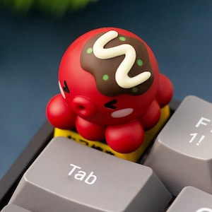 May include: A red octopus-shaped keycap with a brown and white topping, sitting on a yellow key. The keycap is on a gray keyboard with the "Tab" key visible. The octopus has a cute design.