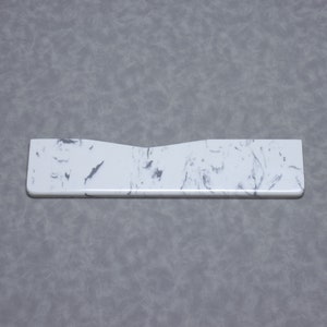 Könnte beinhalten: Weißer Marmorsockel mit wellenförmigem Rand. Der Sockel ist aus glattem, glänzendem Material gefertigt und hat eine dezente Marmorierung.