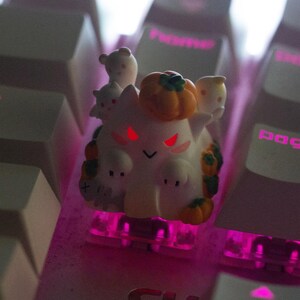 May include: A custom keycap featuring a white ghost-like character with red eyes, a pumpkin on its head, and small ghost companions. The keycap is on a keyboard with pink backlighting, creating a Halloween theme.