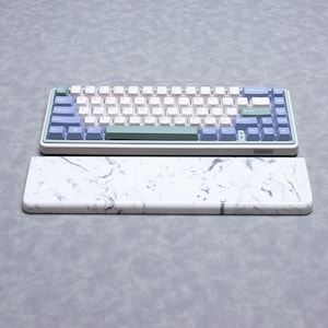 May include: A white and green keyboard with a marble-patterned wrist rest. The keyboard has a light blue and green colour scheme and the wrist rest is white with grey marble-like swirls. The keyboard has the word "NUMILO" printed on the bottom right corner.