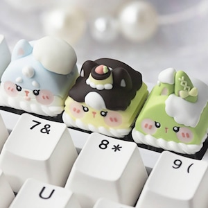 May include: Close-up of three colorful, decorative keyboard keycaps. The keycaps feature cartoon-style designs of animals and cakes, with pastel colors and cute facial expressions. The keys are on a white keyboard.