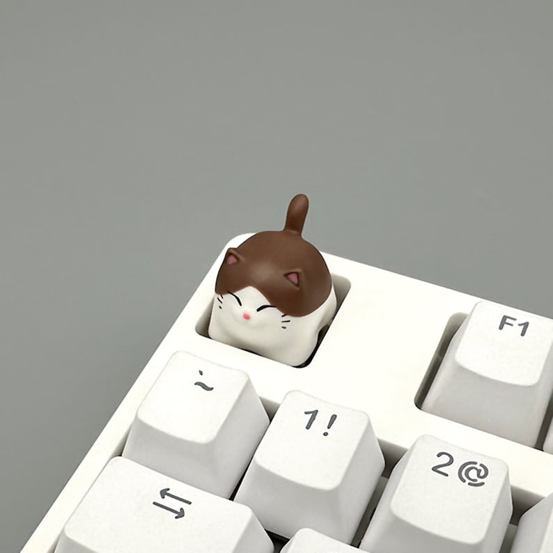 May include: A white keyboard with a brown and white cat keycap on the '1' key. The cat keycap is smiling and has brown ears and a white belly.