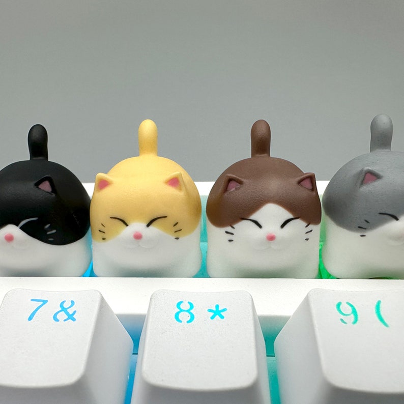 May include: Four cat-shaped keycaps for a mechanical keyboard. The keycaps are black, yellow, brown, and gray. The keycaps are designed to look like cute cartoon cats with white faces and black eyes.
