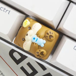 May include: A brown otter-shaped mechanical keyboard keycap featuring a cream-colored face, a small yellow shell on its head, and a light blue figure held in its paws. The keycap is positioned between white keyboard keys with the text ctrl visible.