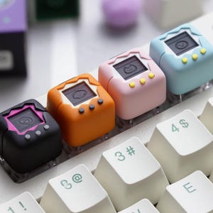 May include: A close-up of a mechanical keyboard with custom keycaps. The keycaps are shaped like retro handheld game consoles in black, orange, pink, and blue. Each keycap has a small screen and buttons. The keyboard keys are white.