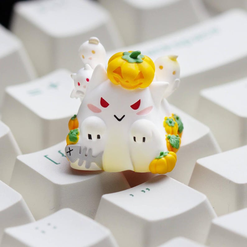 May include: A white and yellow Halloween-themed keyboard keycap featuring a ghost character with a jack-o'-lantern on its head. The keycap has red eyes, pink cheeks, and small pumpkins around its base. The keycap is resting on a white keyboard.
