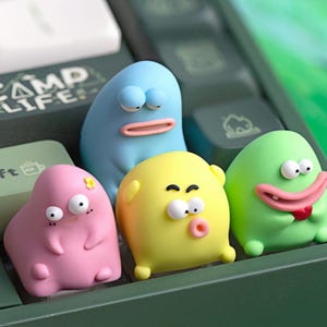 May include: Four colorful, cartoonish figures with expressive faces rest on a green surface. The figures are in shades of pink, yellow, blue, and green. The blue figure has a wide mouth, while the green figure has a large, smiling mouth. The word "CAMP LIFE" is visible.