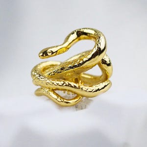 May include: A gold-toned ring shaped like a coiled snake. The metal surface features a textured, scale-like pattern throughout the body of the serpent. The snake's head is positioned at the top, while the tail curves around the bottom of the band.