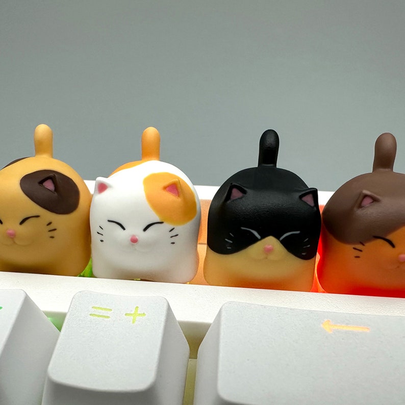 May include: Four colorful cat-shaped keycaps for a keyboard. The keycaps are orange, white, black, and brown. The keycaps are sitting on a white keyboard.