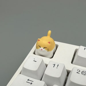 May include: A yellow and white cat-shaped keycap with a smiling face. The keycap is sitting on a white keyboard.