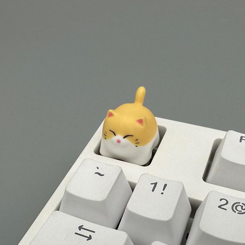 May include: A yellow and white cat-shaped keycap with a smiling face. The keycap is sitting on a white keyboard.
