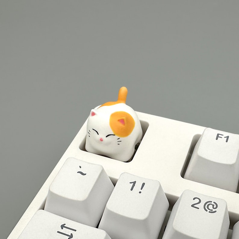 May include: A white and orange cat keycap with a smiling face, sitting on a white keyboard with keys labeled "~", "1!", "2@", and "F1".