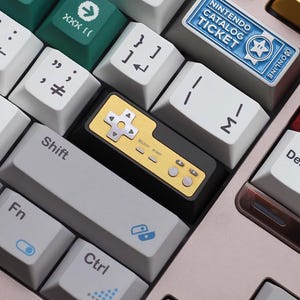 Puede incluir: Primer plano de un teclado mecánico con teclas personalizadas de varios colores. Una tecla dorada con forma de mando de juego retro está en el centro. El teclado tiene las palabras "Shift", "Fn" y "Ctrl".